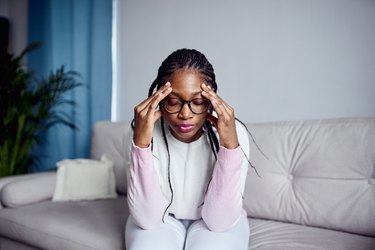 Person with braids and glasses wearing a pink sweater sitting on the couch and holding their head to keep themselves from getting a silent migraine