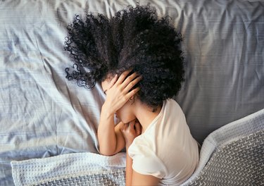 a person with dark brown curly hair lying in bed with their hand over their ear because they have sharp pain in the ear