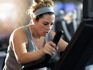 Person in a tank top with a blonde bun and hair band doing a 20-minute low-impact elliptical HIIT workout at the gym