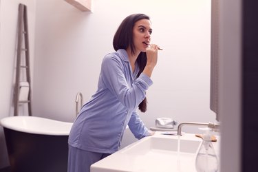 woman wearing pajamas and brushing her teeth in front of the bathroom mirror