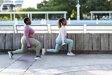 Two people doing forward lunges outside in a city park.
