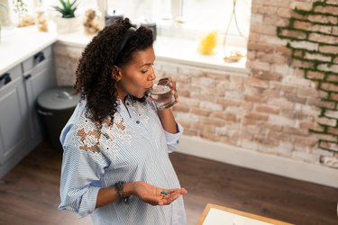 Pregnant lady in her kitchen taking her prenatal vitamins with a glass of water