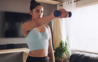 Close-up of person wearing a blue sports bra and black leggings working out with dumbbells in living room.