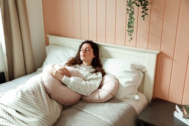 a person with long brown curly hair lying in bed holding a pink pillow over their stomach because they're feeling nauseous on their period