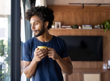 A person with curly brown hair wearing a navy blue t-shirt standing in a kitchen drinking a cup of coffee while staring out a window