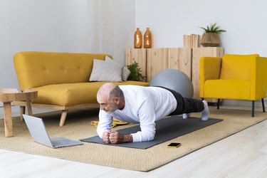 Older adult with laptop performing plank pose on exercise mat in living room to demonstrate a body-weight workout for older adults.