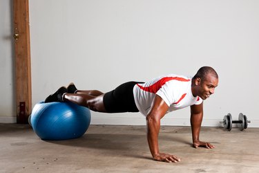 Man in a white shirt with red stripes and black shorts with feet balancing on a blue stability ball, performing a plank during a stability ball workout at home